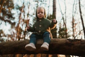 Child sits on a log holding a dried leaf, surrounded by bare trees, capturing curiosity, stillness, and seasonal change