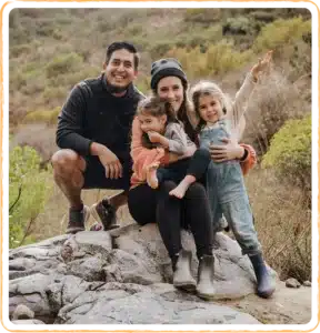  A family with parents and two daughters sitting together on a rock outdoors, smiling and holding each other