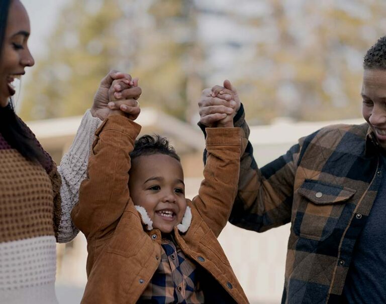 A smiling young child is lifted up by two adults holding their hands outdoors.