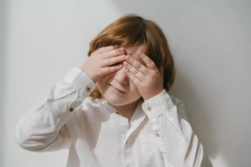 A young child with reddish-brown hair wearing a white button-up shirt, covering their eyes with both hands against a light background.