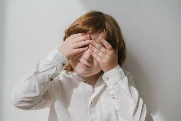 A young child with reddish-brown hair wearing a white button-up shirt, covering their eyes with both hands against a light background.