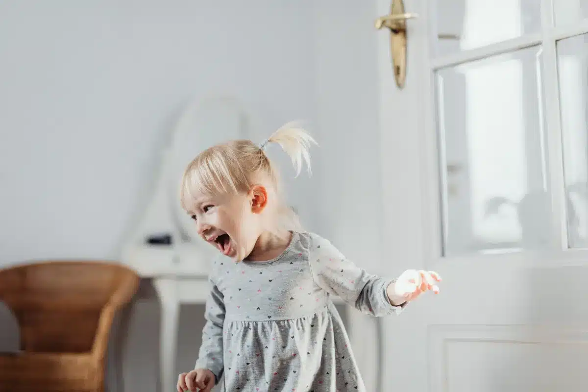 A joyful toddler girl with blonde hair in a ponytail laughing excitedly while playing indoors near a white door.