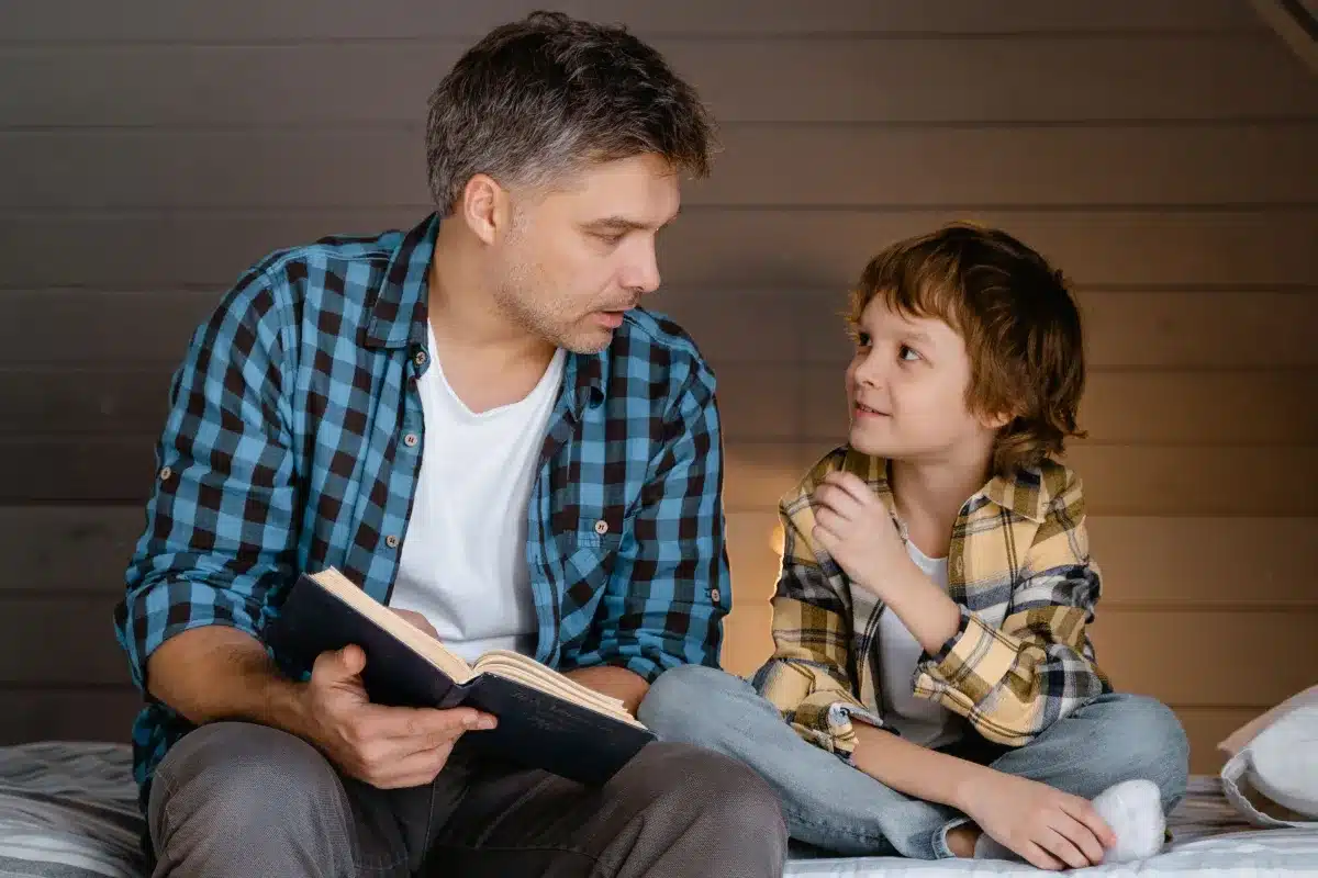 A father in a blue flannel shirt holding an open book and talking to his young son, who is sitting cross-legged on a bed wearing a yellow plaid shirt and listening intently.