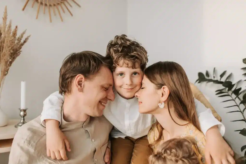 A happy family sharing a close embrace indoors, featuring a mother and father smiling warmly next to their young son with curly hair.