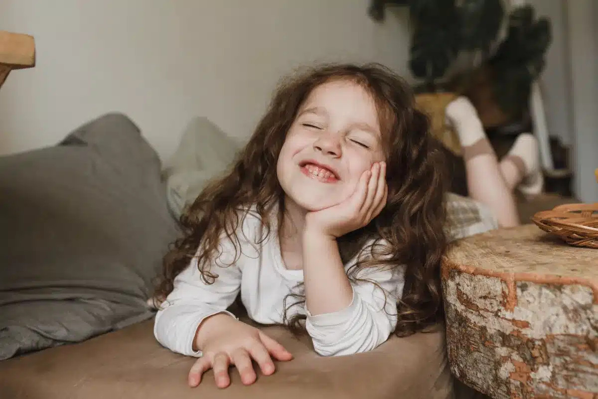 A happy young girl with long, curly brown hair lying on a brown couch, smiling with her eyes closed and her chin resting on her hand.
