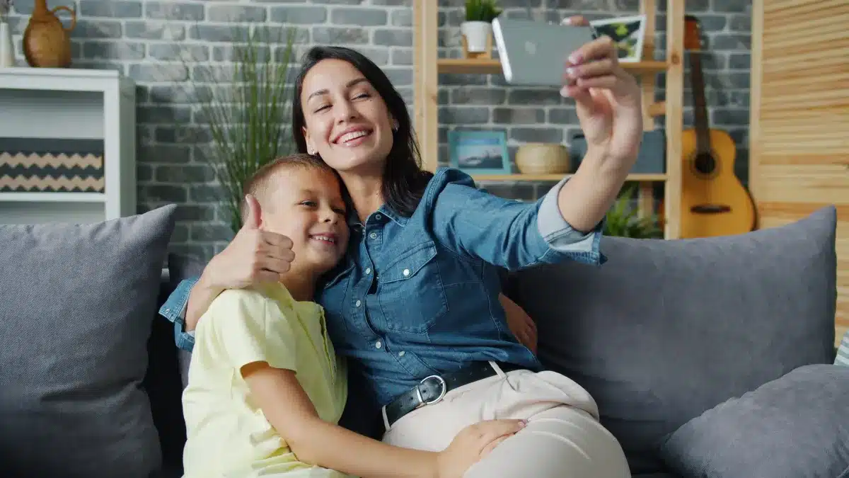 A smiling mother in a denim shirt holds up a smartphone to take a selfie on a grey couch, while hugging her young son who is smiling and giving a thumbs up.